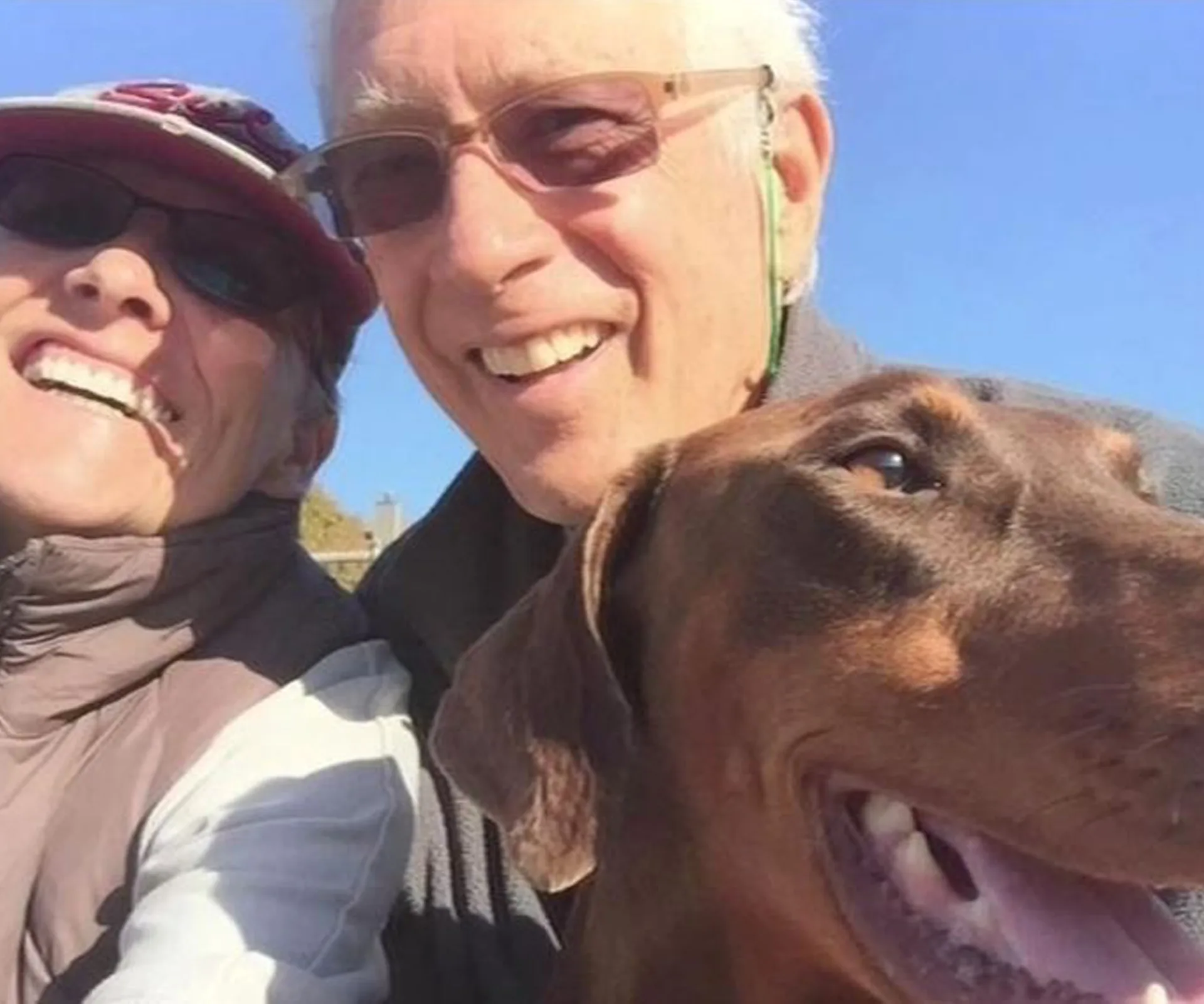 Elderly couple smiling with a brown dog outdoors under clear blue sky.
