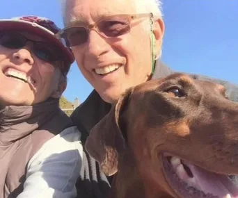 Elderly couple smiling with a brown dog outdoors under clear blue sky.