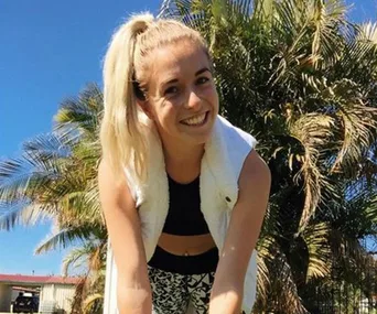 A smiling woman with a ponytail outdoors, wearing a black sports bra and patterned leggings, with palm trees in the background.