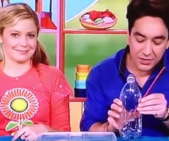 Two presenters from a children's show demonstrate a craft using a plastic bottle and straw at a colorful table.