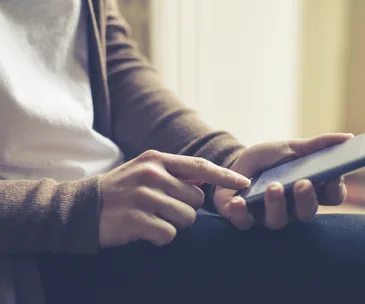 Person using a smartphone, pointing at the screen with one hand, wearing a white shirt and brown cardigan.