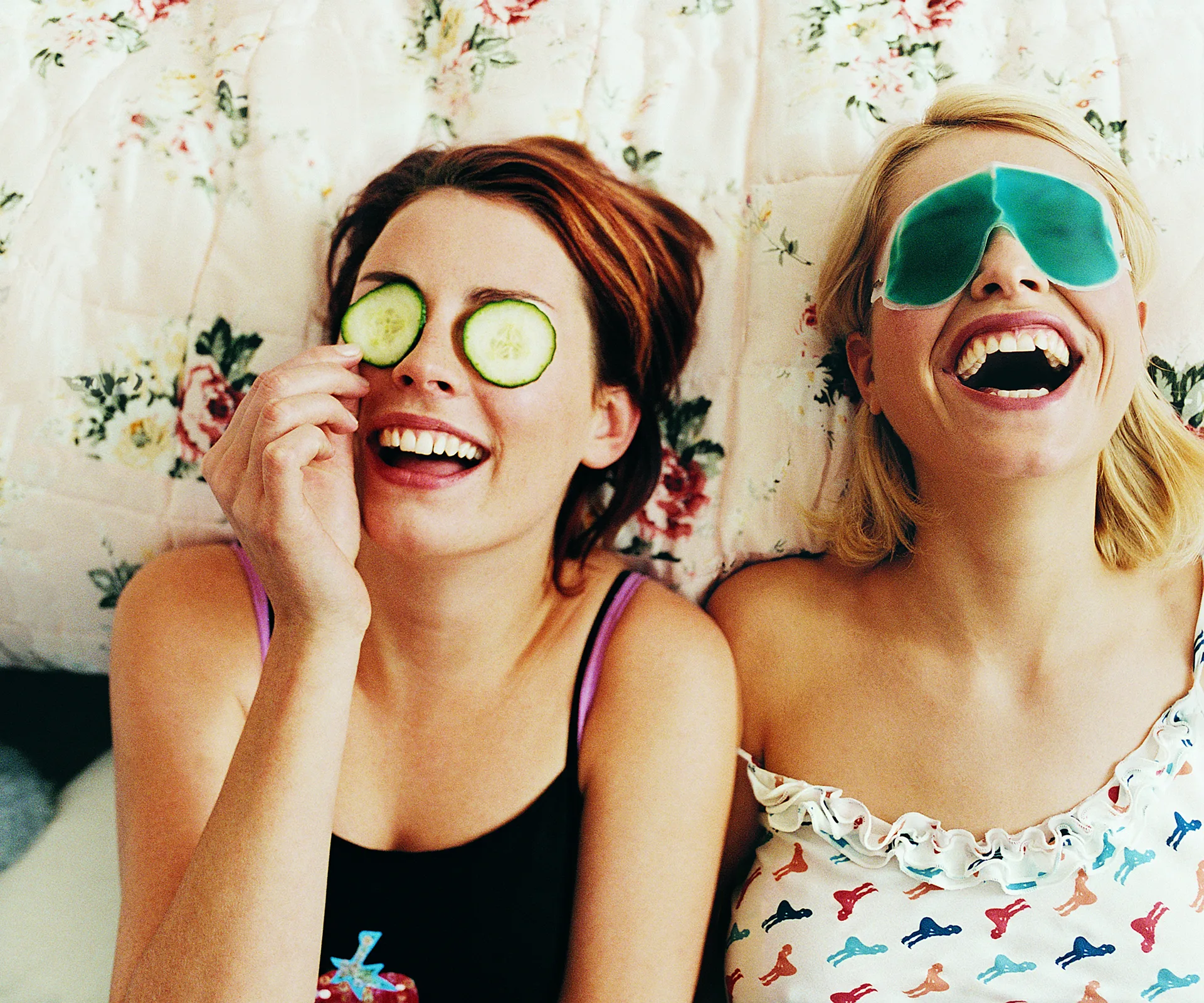 Two women laughing, lying on a floral bedspread; one with cucumber slices on her eyes, the other with a gel eye mask.