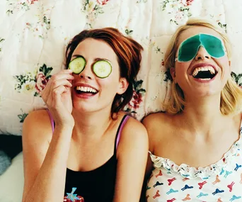 Two women laughing, lying on a floral bedspread; one with cucumber slices on her eyes, the other with a gel eye mask.