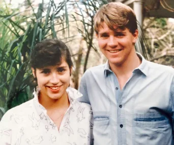 A smiling young man and woman standing close together outdoors with greenery in the background.