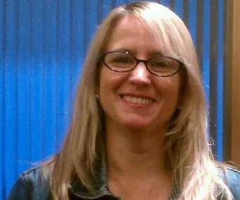 A woman with glasses and blonde hair smiling against a blue background.