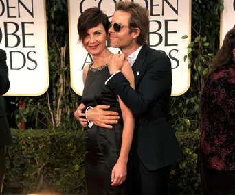 A man in sunglasses embraces a woman in a black dress at an awards event, smiling for the camera.