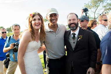 Wedding couple poses with a smiling man in a baseball cap, surrounded by photographers.