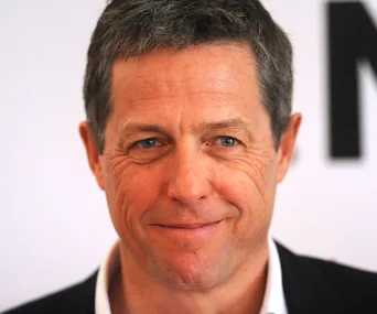 A person smiling at an event or press gathering, wearing a suit and standing against a plain background.