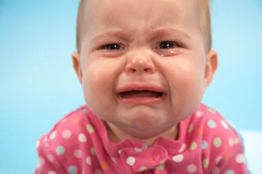 A crying baby wearing a pink polka dot onesie with a blue background.