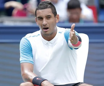 Tennis player gesturing on court with an expressive face, wearing a white and blue shirt.