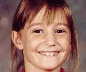 A young girl with long brown hair and bangs smiling at the camera in a school photo.