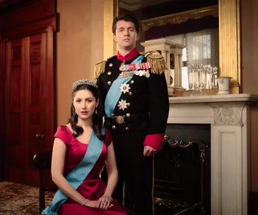 A woman in a royal dress and tiara seated, with a man in formal uniform standing beside her in an opulent room.