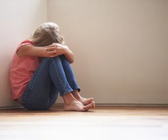 A young girl sits curled up against a wall, head on arms, conveying sadness and vulnerability.