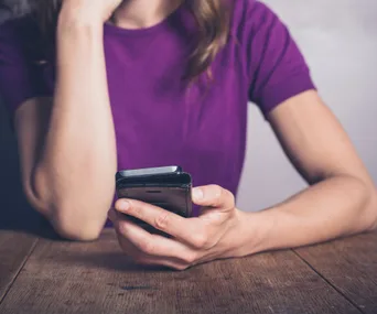 A woman in a purple shirt sits at a wooden table, holding and looking at her smartphone.