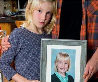 Young girl holding a framed school photo of herself, looking upset while a hand rests on her shoulder.