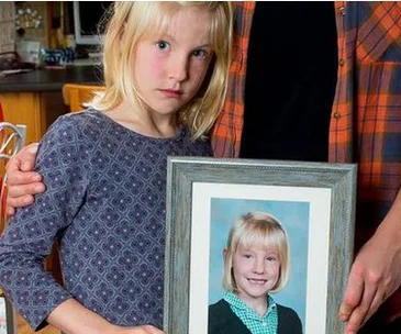 Young girl holding a framed school photo of herself, looking upset while a hand rests on her shoulder.