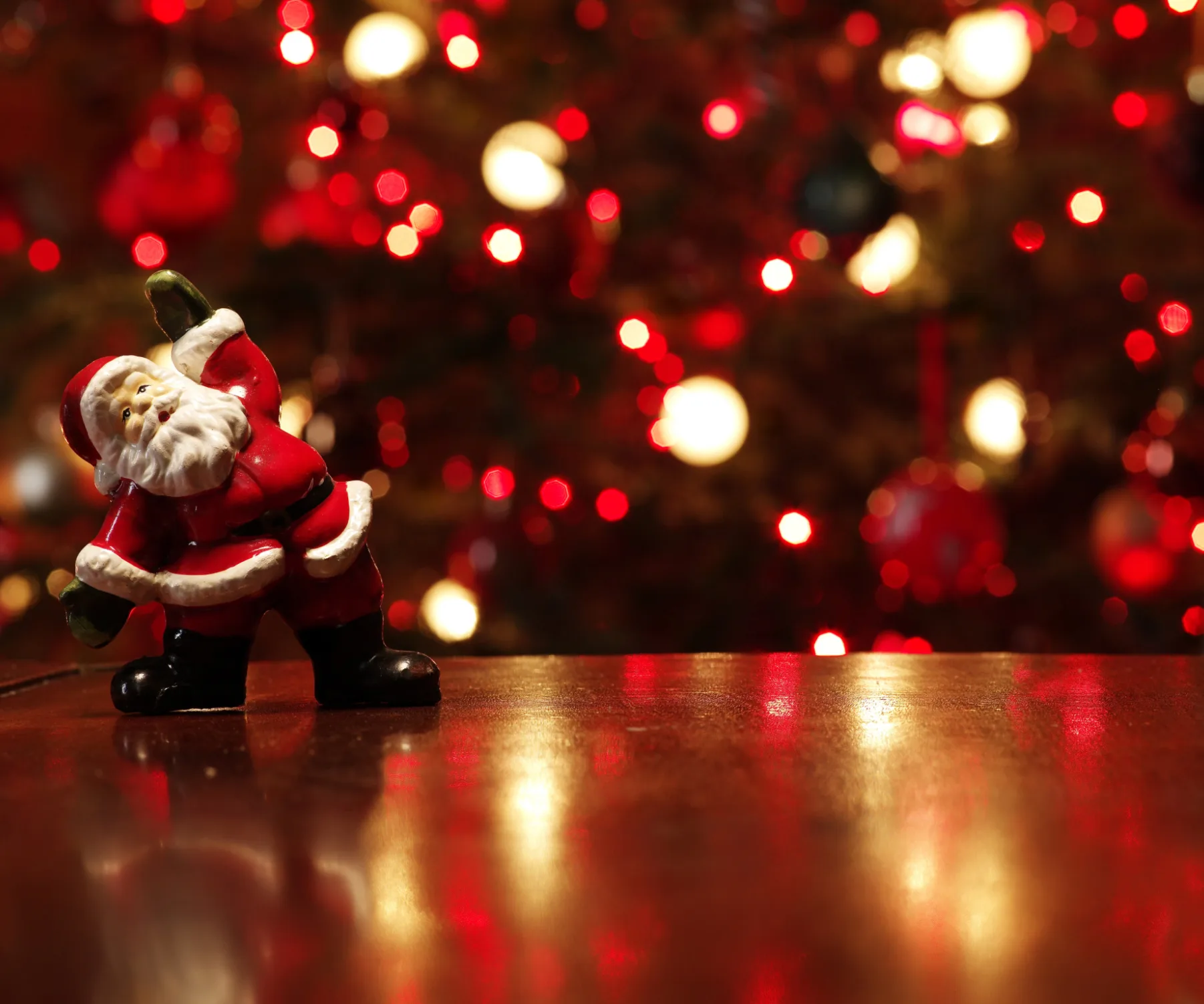 A small Santa Claus figure on a wooden surface with a blurred, glowing red Christmas background.