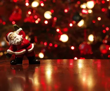 A small Santa Claus figure on a wooden surface with a blurred, glowing red Christmas background.