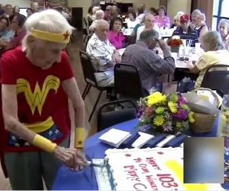 103-year-old woman cutting her birthday cake dressed as Wonder Woman, surrounded by guests at a celebration.