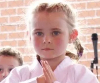 A young girl in a karate uniform poses with hands together, a brick wall in the background.