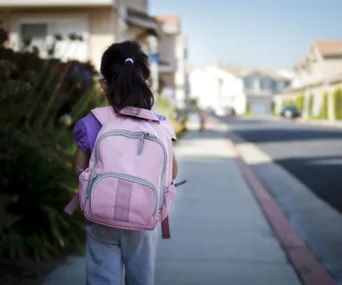 A young girl with a ponytail, wearing a pink backpack, walks on a suburban sidewalk in the morning.