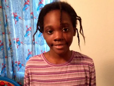Young girl in a striped shirt stands in front of Spider-Man curtains, smiling gently with loose, twisted hair strands.