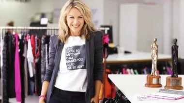Woman in a casual outfit smiles in a chic clothing store, with stylish racks and trophies visible on a table.