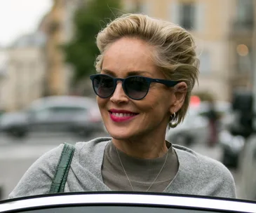 A woman with short blonde hair, wearing sunglasses and a grey top, smiles outdoors with a blurry urban background.