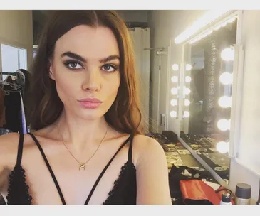 A woman taking a selfie in a dressing room with bright lights and a vanity mirror in the background.