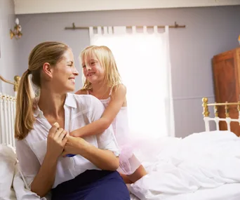 Mother and daughter smiling and hugging on a bed in a sunlit bedroom.