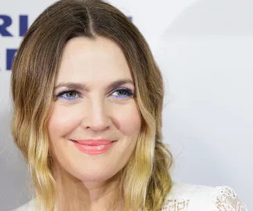 Drew Barrymore smiles at an event, wearing light makeup and a white lace top.