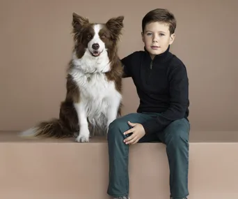 Boy in a dark sweater sits next to a brown and white dog, both facing forward against a beige background.