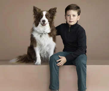 Boy in a dark sweater sits next to a brown and white dog, both facing forward against a beige background.