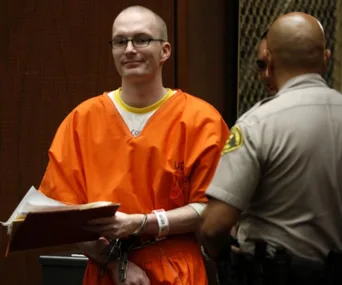 A man in an orange prison jumpsuit stands in a courtroom, holding papers, with handcuffs on his wrists.