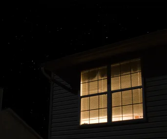 Upper floor window of house at night, lights on inside against a starry sky.