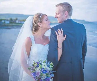Bride in white dress and groom in suit embrace on a beach, with a bouquet of flowers and ocean in the background.