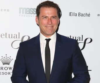 Man in a dark suit smiling at a formal event with branded wall in background.