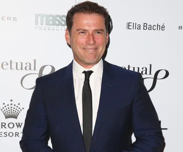 Man in a dark suit smiling at a formal event with branded wall in background.