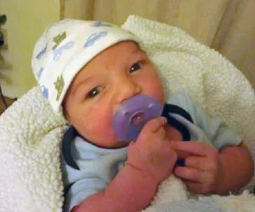 A baby wearing a white hat with car patterns, holding a purple pacifier, and wrapped in a soft white blanket.