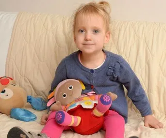 A young girl with blond hair sits on a sofa holding a plush doll, with another toy beside her.