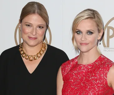 Two women posing together at an event, one in a black top with a gold necklace, the other in a red lace dress.