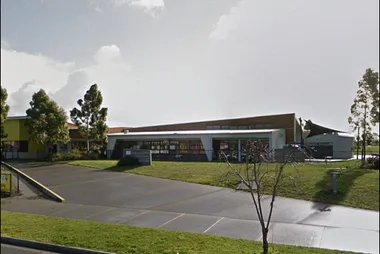 School building with modern architecture, surrounded by trees and a grassy area under a cloudy sky.