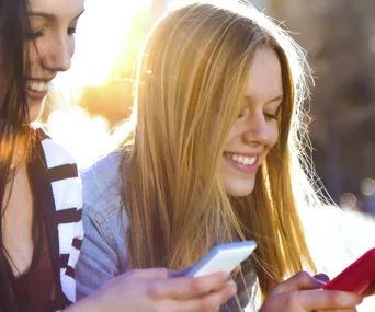Two smiling young women looking at their phones with sunlight in the background.