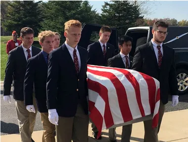 Teenage pallbearers carry a coffin draped with the American flag at a funeral service outdoors.