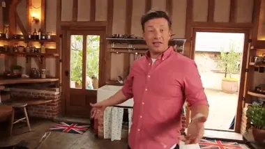 Jamie Oliver in a kitchen, gesturing while filming, with a rustic backdrop and British-themed mats on the floor.