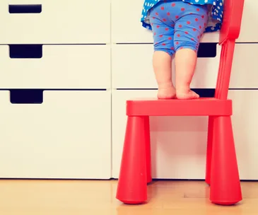 Child standing on a red chair reaching a white dresser, indicating a potential safety hazard.