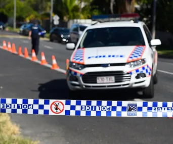 Police car behind tape with "POLICE" and no entry symbol. Cones set up on road. Blurred officer walking.