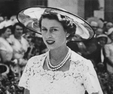 A woman in a lace dress and hat, wearing a pearl necklace, smiles in a crowd.