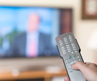 Hand holding a remote control in front of a blurred television screen.