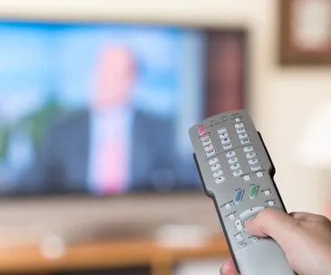 Hand holding a remote control in front of a blurred television screen.